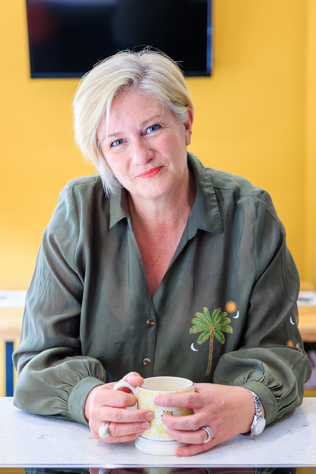 Photo of Karen A woman with short blonde hair sits at a table, holding a mug, against a yellow background.